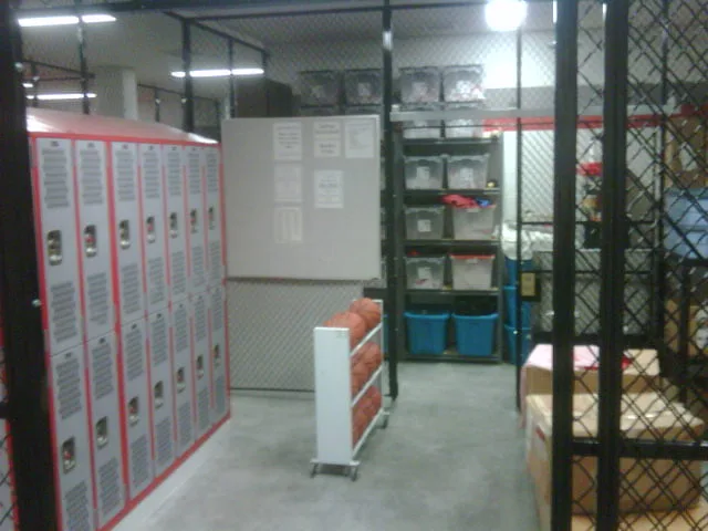 A basketball locker room with red-trimmed lockers, a rack of basketballs and shelves of gear bins